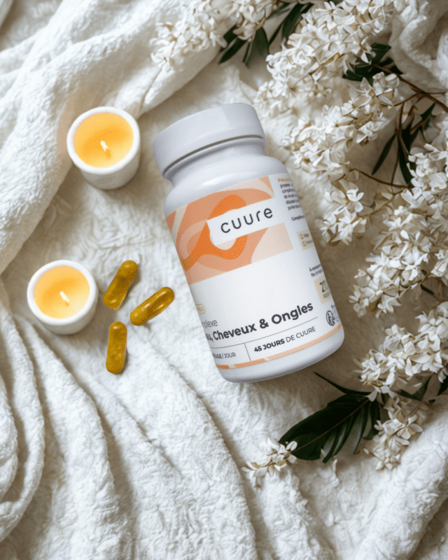 Pot du complément alimentaire Cuure peau, cheveux et ongles posé sur une serviette de salle de bain avec gélules jaunes, bougie et fleurs blanches. Ambiance spa bien-être